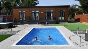 A rectangular swimming pool with two people in it, surrounded by a deck area with a wooden building in the background. - Spanhoe Lodge - B&B (Corby)