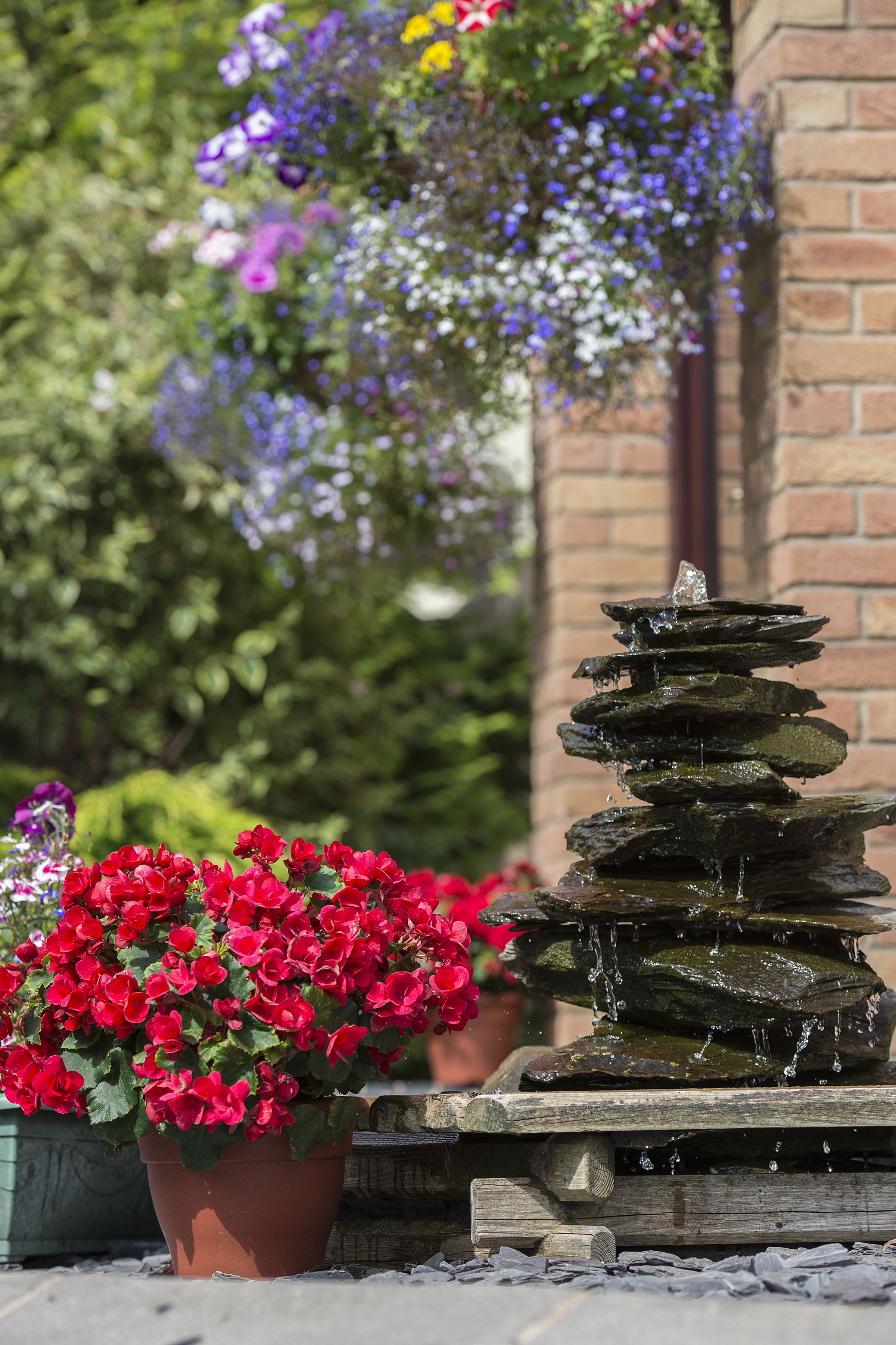 Una fuente de piedra escalonada con agua cayendo en cascada, rodeada de flores rojas y moradas en un jardín.