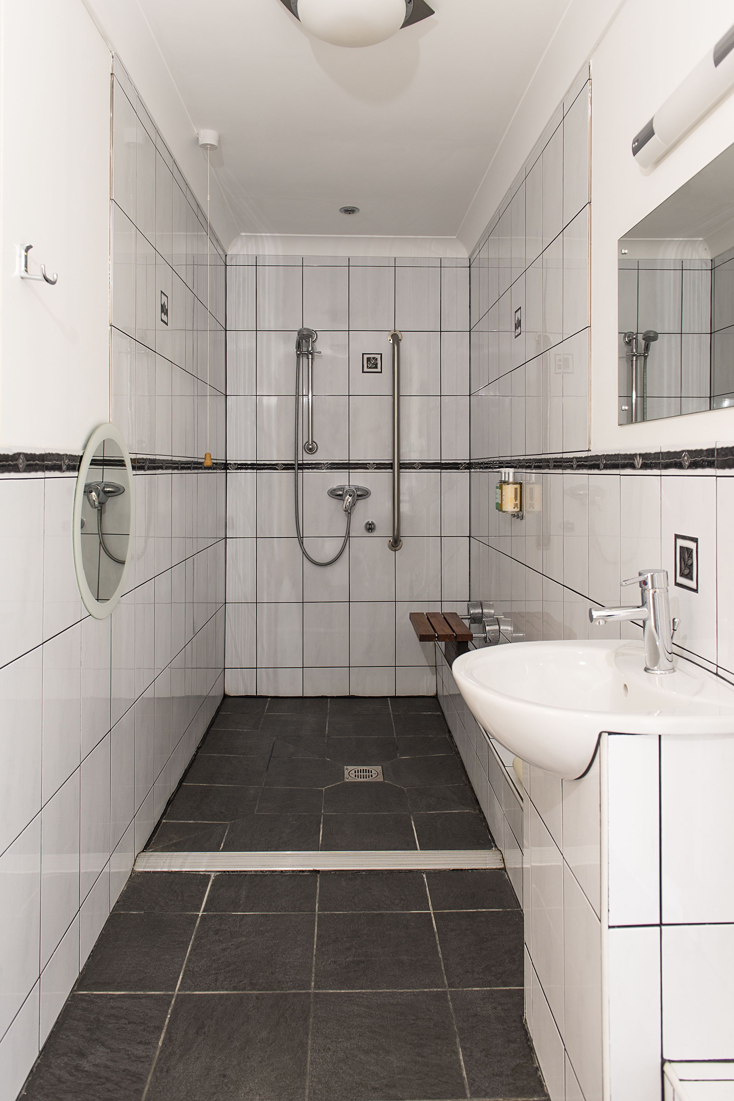 A bathroom with white tiled walls, a black tiled floor, a white sink, a mirror, and a shower area.