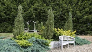 A garden with a white bench, tall coniferous trees, and a wooden pergola. - Spanhoe Lodge - B&B (Corby)