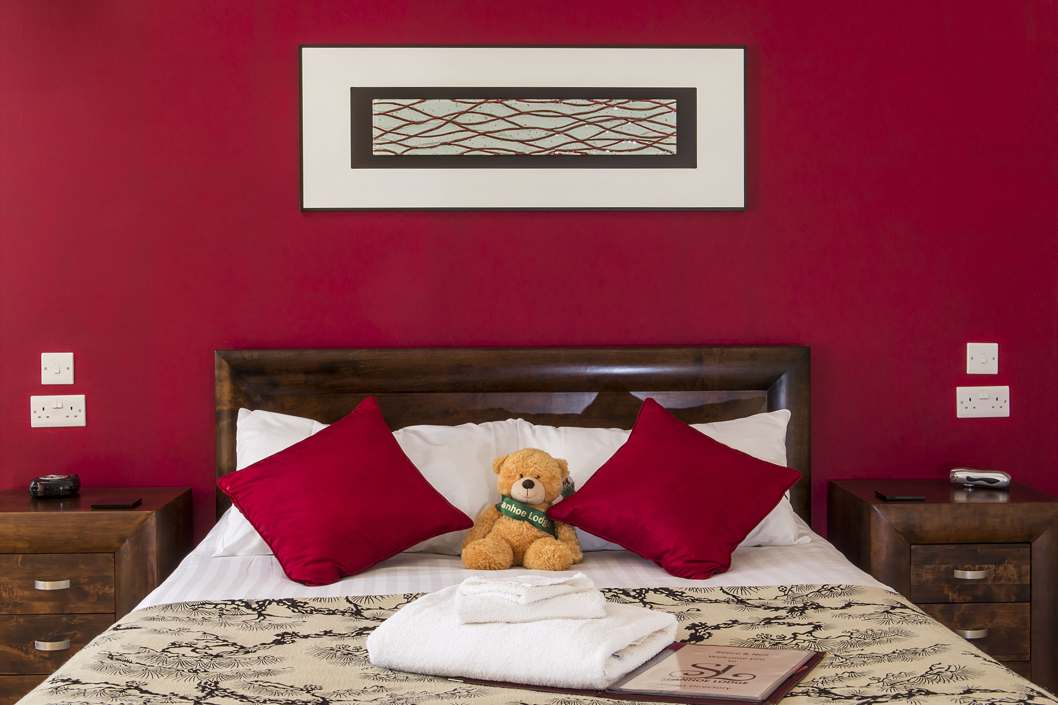Una cama bien hecha con un osito de peluche, almohadas rojas y una obra de arte enmarcada encima.