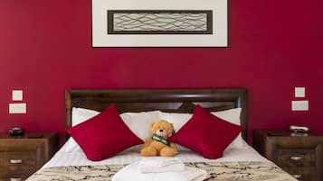 Una cama bien hecha con un osito de peluche, almohadas rojas y una obra de arte enmarcada encima.