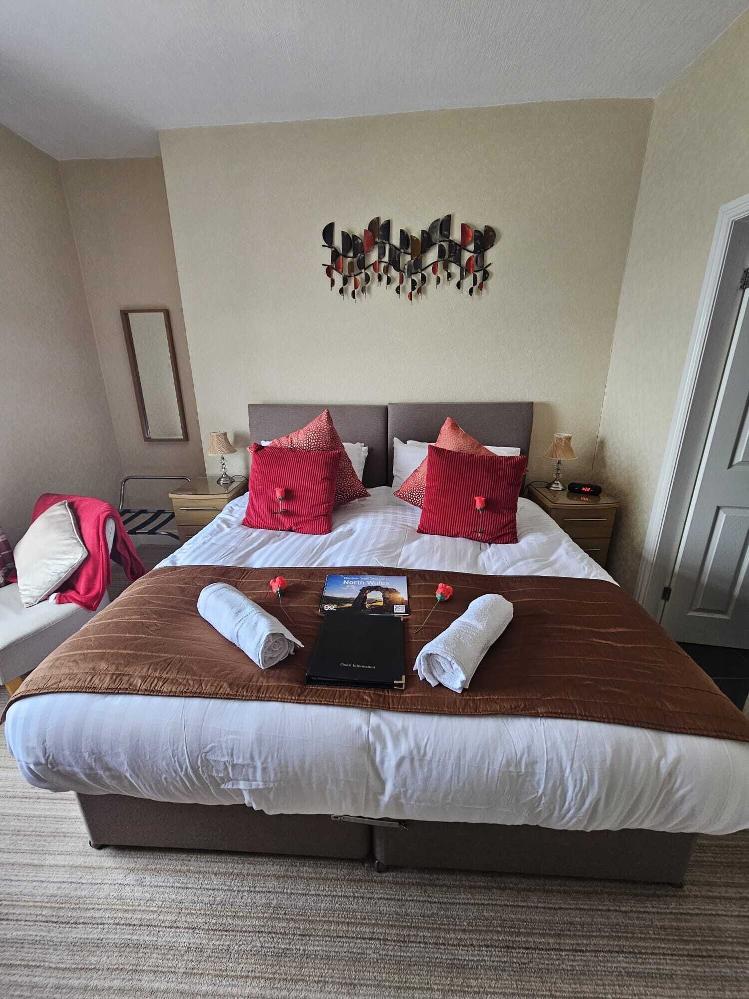 A hotel room with a large bed, two red pillows, a decorative wall piece, a nightstand with a lamp, and a book titled "North Wales".