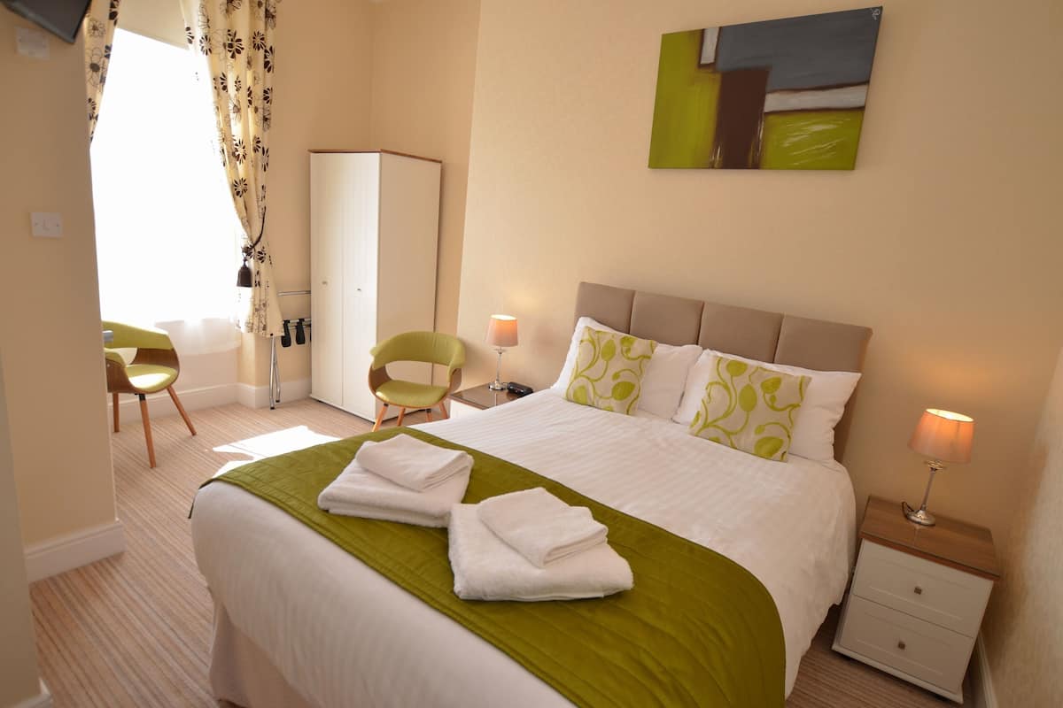 A neatly made bed with a green bedspread and white pillows, a bedside lamp, and a chair in a well-lit room with a large window and a painting on the wall.