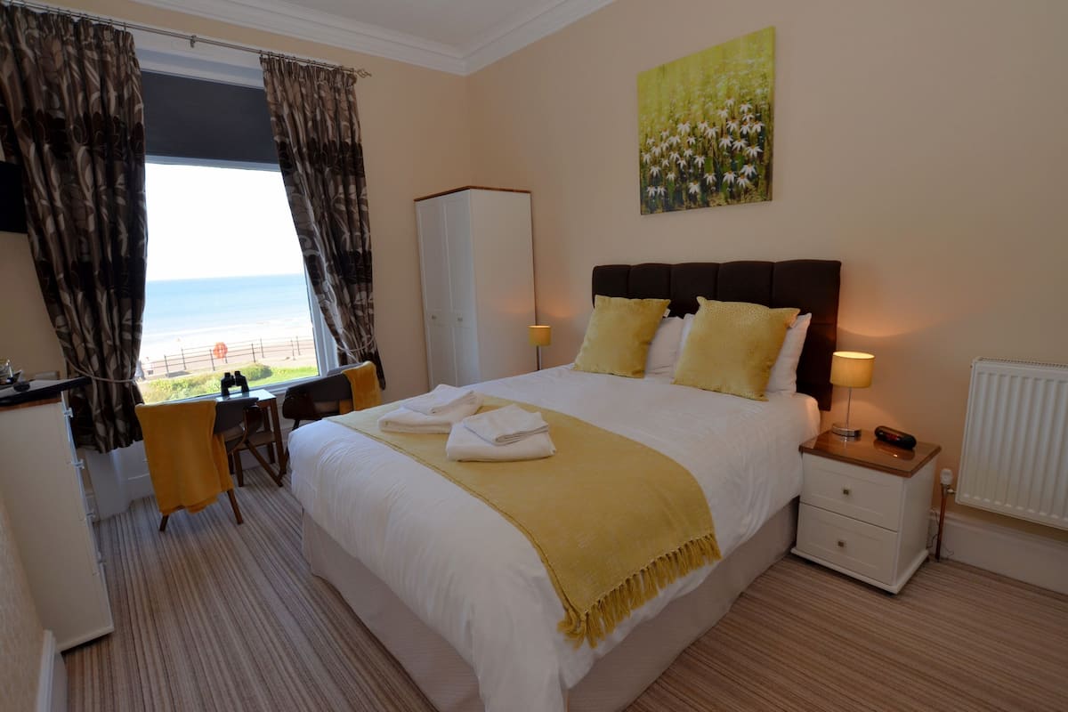 A hotel room with a bed, a desk, a chair, a wardrobe, and a view of the beach.