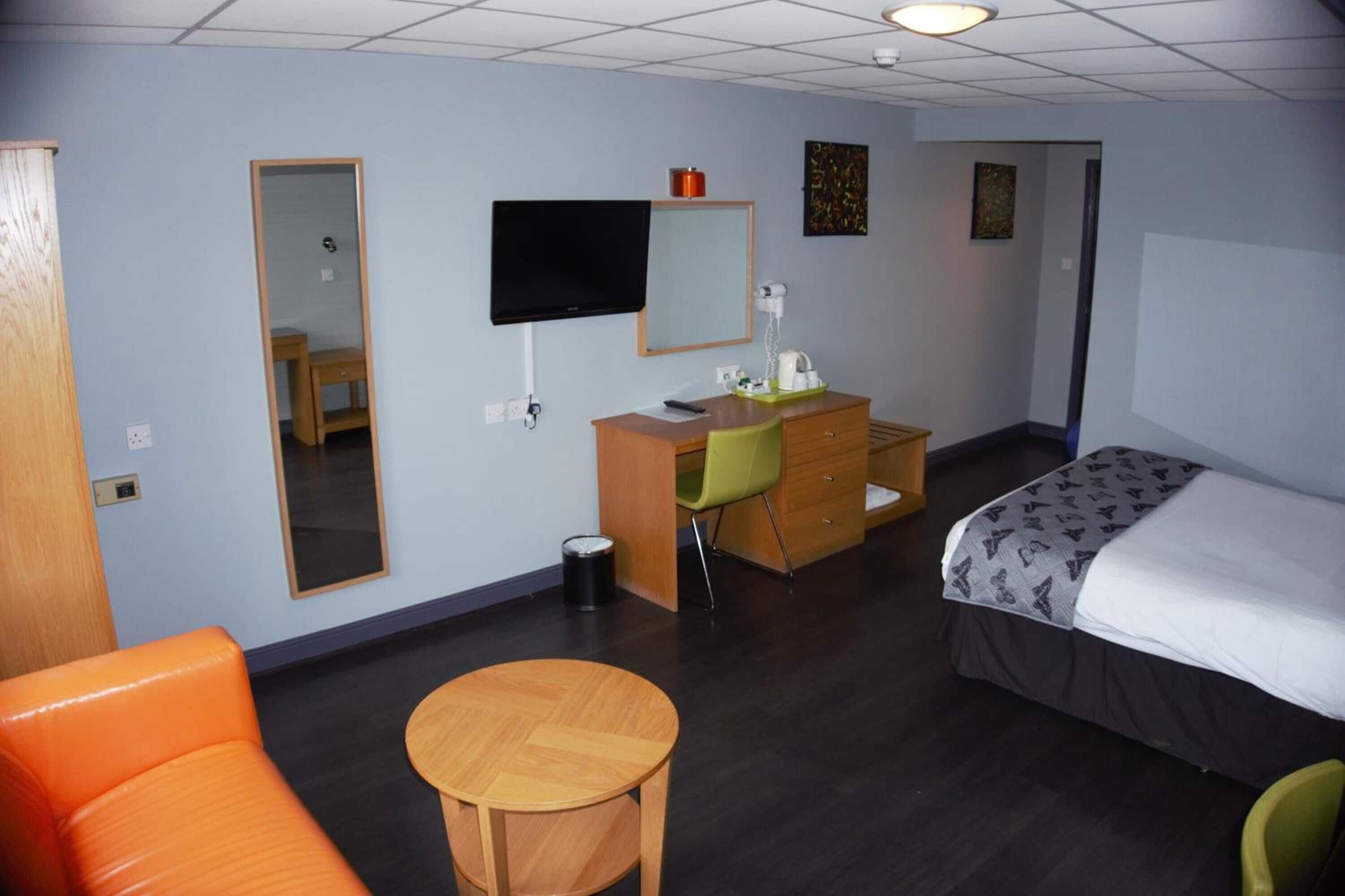 A hotel room with a bed, a desk, a chair, a small table, a mirror, and a television.