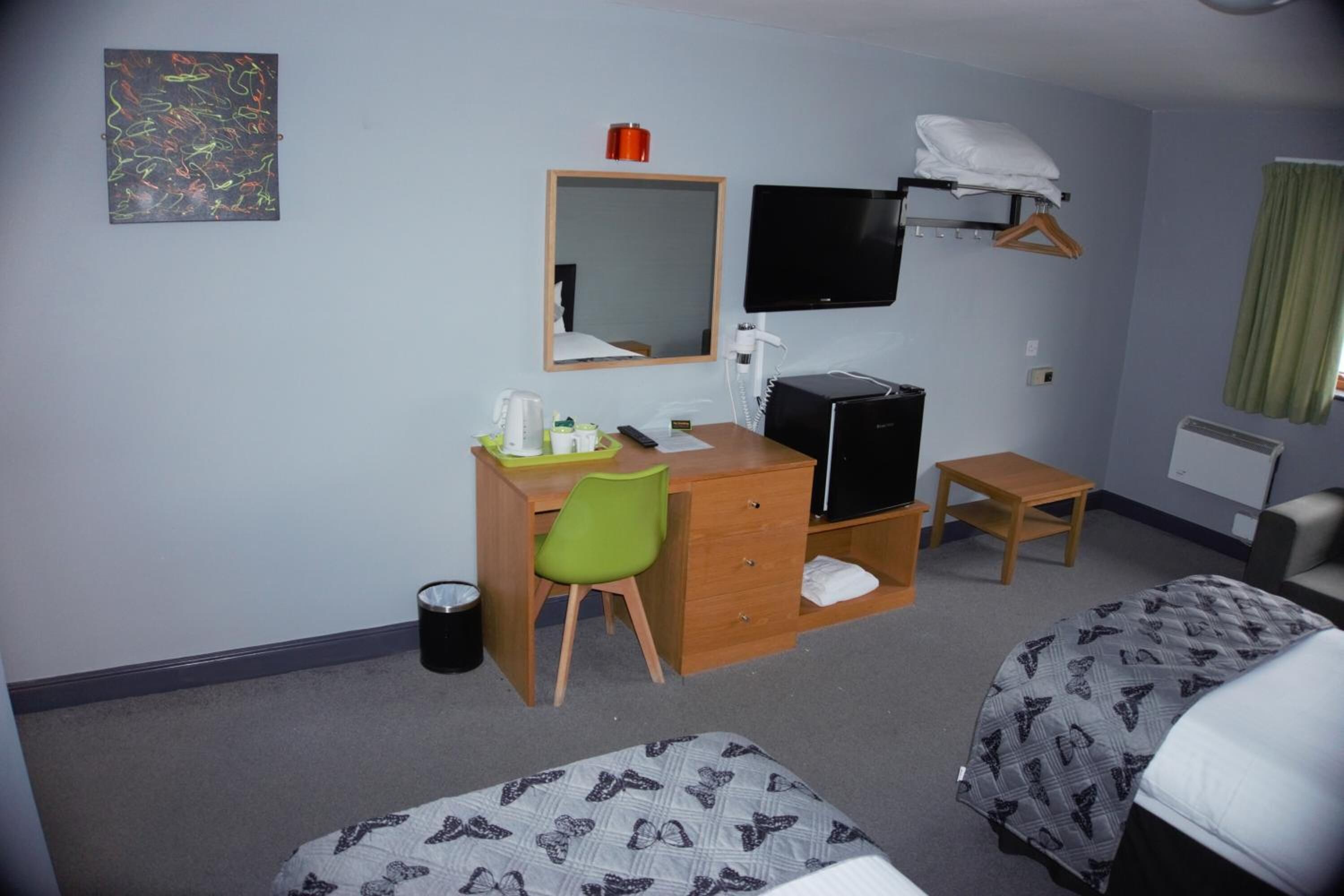 A hotel room with a desk, chair, two beds, a TV, a mirror, and a small table.