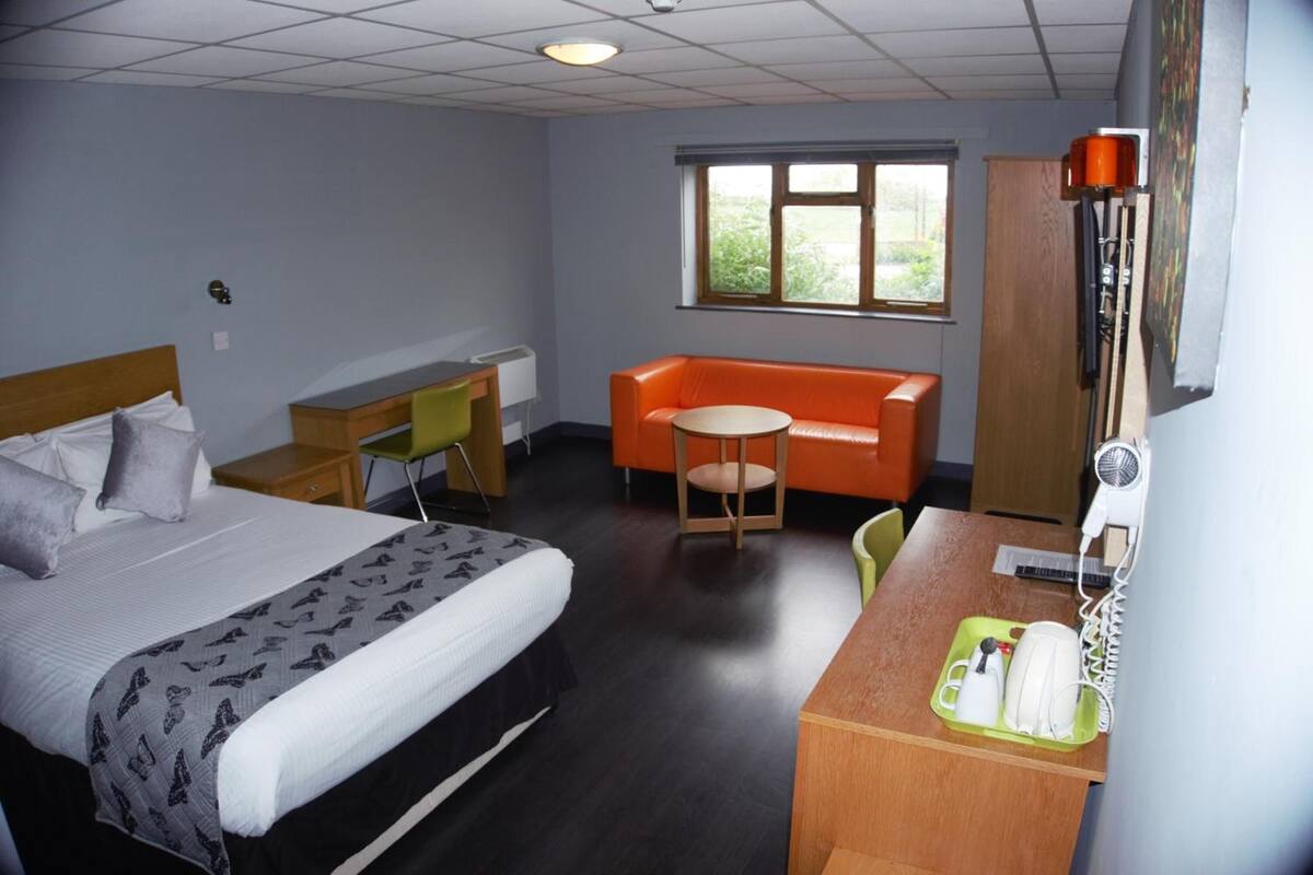 A hotel room with a bed, a desk with a telephone and a hairdryer, an orange sofa, and a window with a view of greenery.