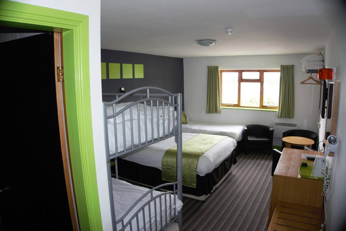 A hotel room with bunk beds, a desk, a chair, a small table, a window with curtains, and a coat rack.