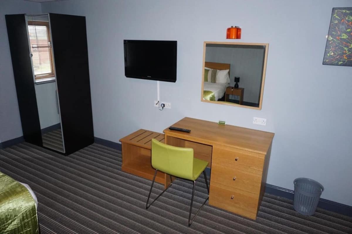 A hotel room with a wooden desk, a green chair, a television, a mirror, and a window with blinds.