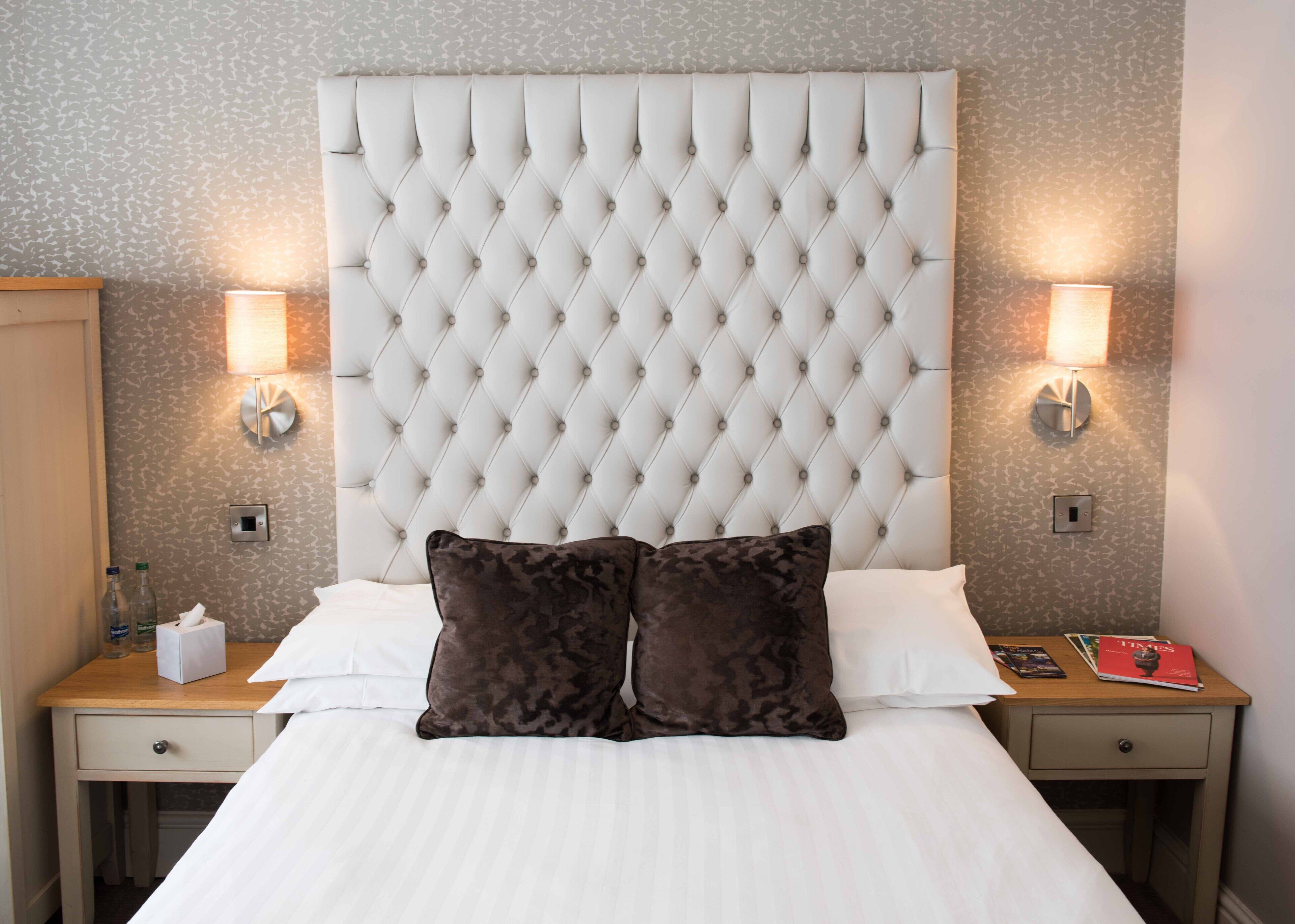 A hotel room with a tufted headboard, two bedside lamps, a wooden nightstand, a bedside table, and a bed with white linens and two dark pillows.