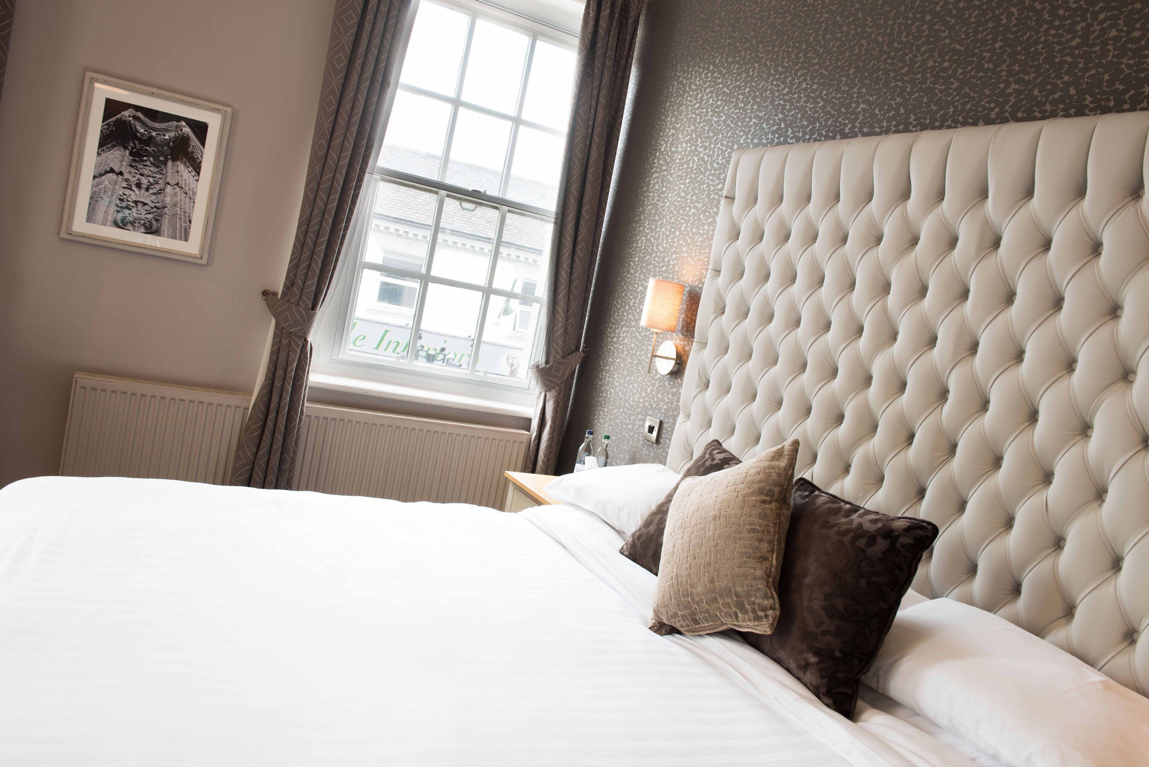 A hotel room with a large bed, a tufted headboard, a framed picture on the wall, a window with curtains, and a nightstand with a lamp.