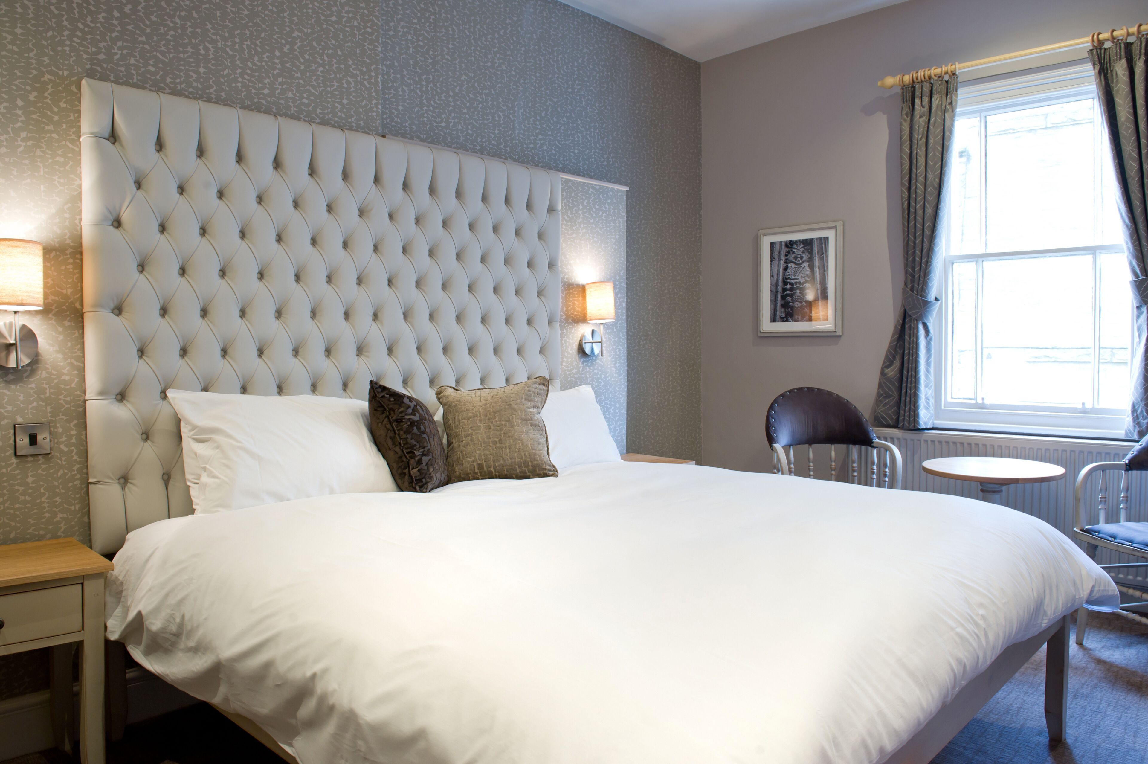 A neatly made bed with a tufted headboard, a bedside table, a chair, and a window with curtains.
