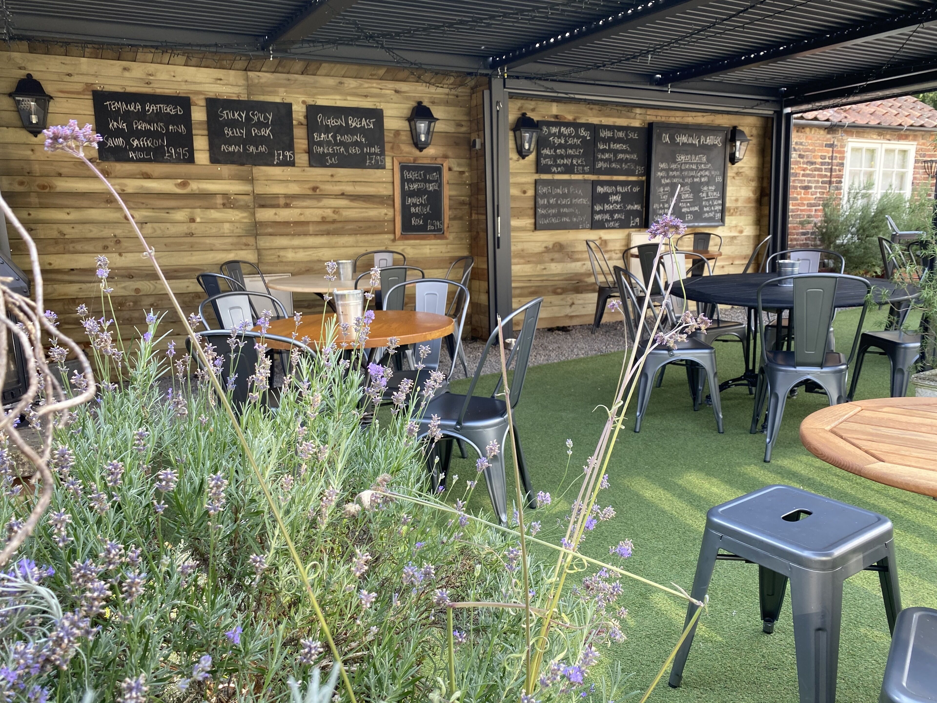 Un comedor al aire libre con paredes de madera y pizarras con menús que incluyen platos como 'Temura Battered King Prawns' y 'Sticky Spicy Belly Pork'.