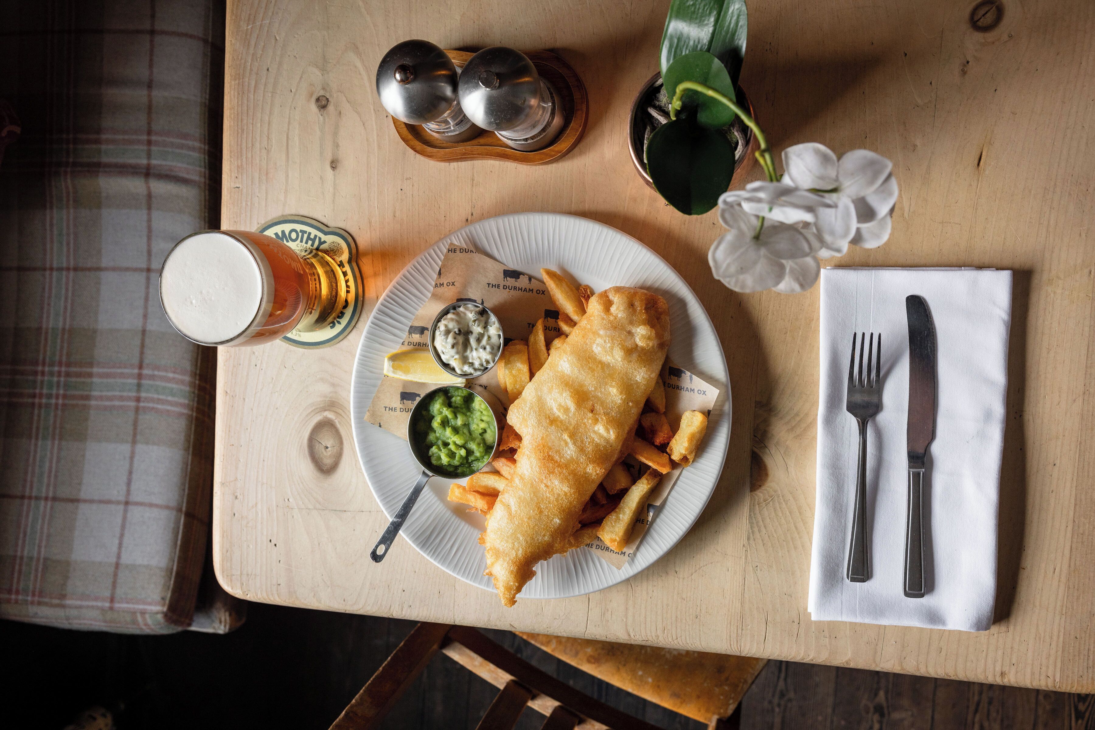 A table with a plate of fish and chips, a glass of beer, condiments, and cutlery.