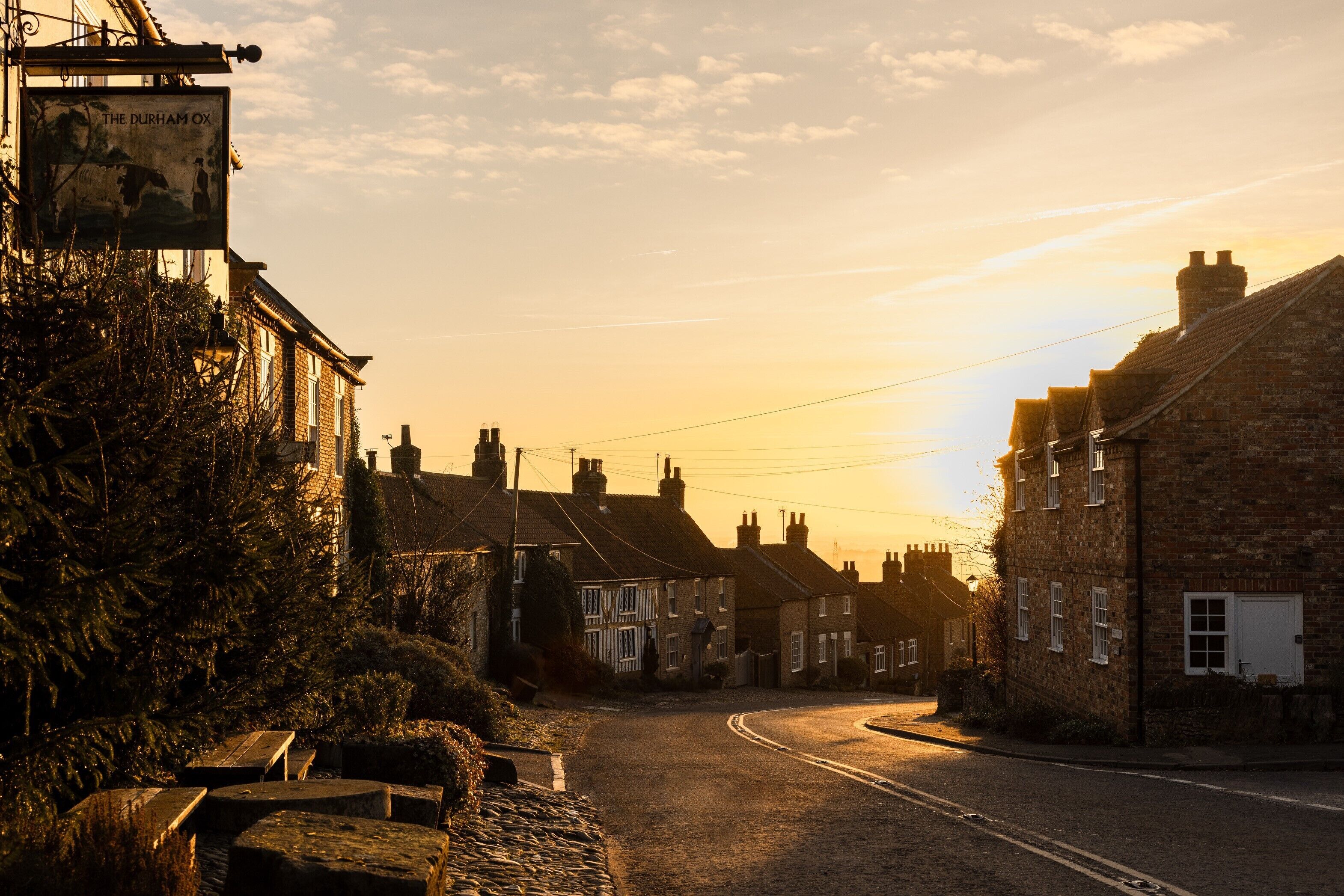 Rua com edifícios tradicionais, um letreiro com 'The Durham Ox' e céu claro.