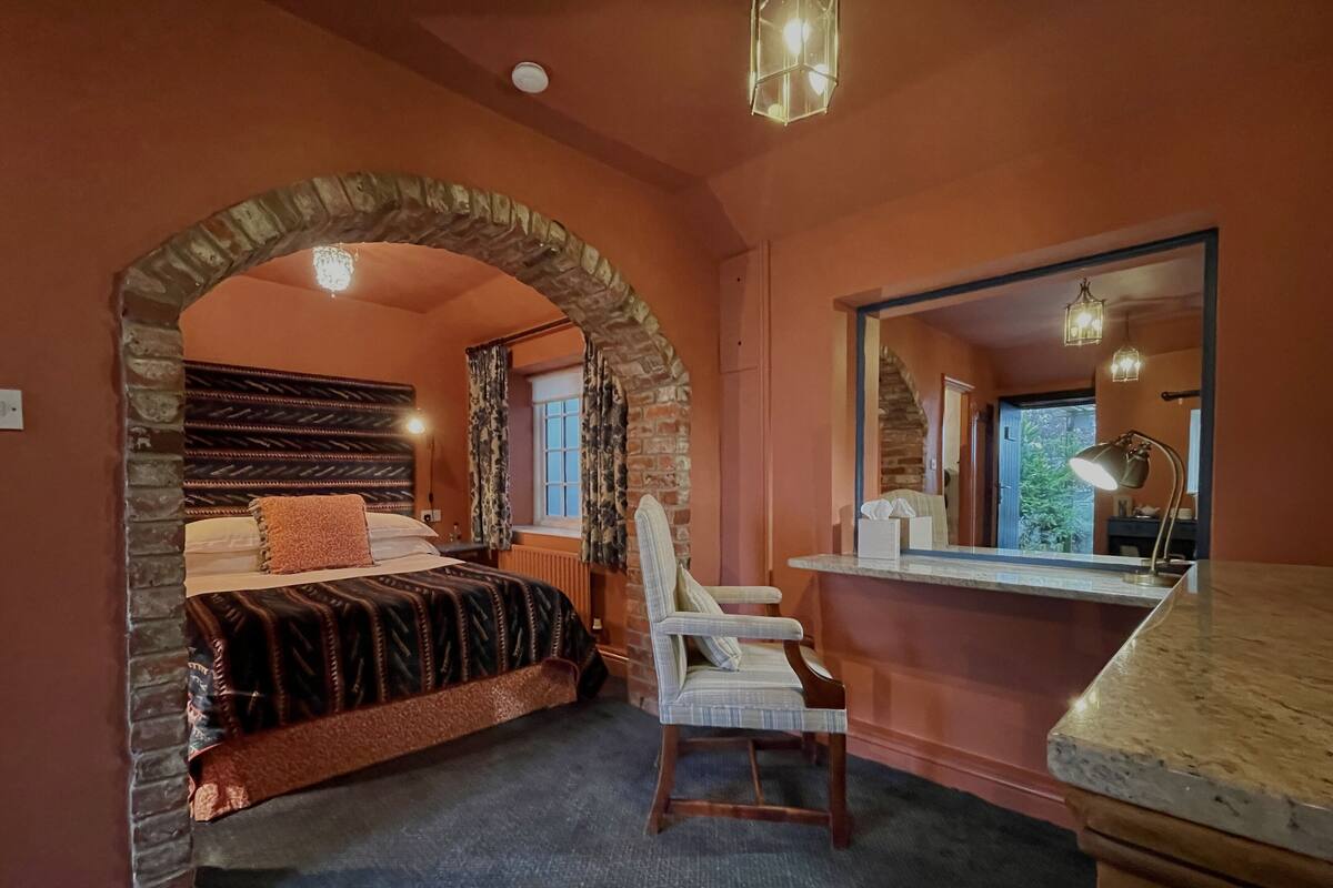 A bedroom with a stone archway, a bed with patterned bedding, a chair, a desk with a mirror, and a view of greenery outside.
