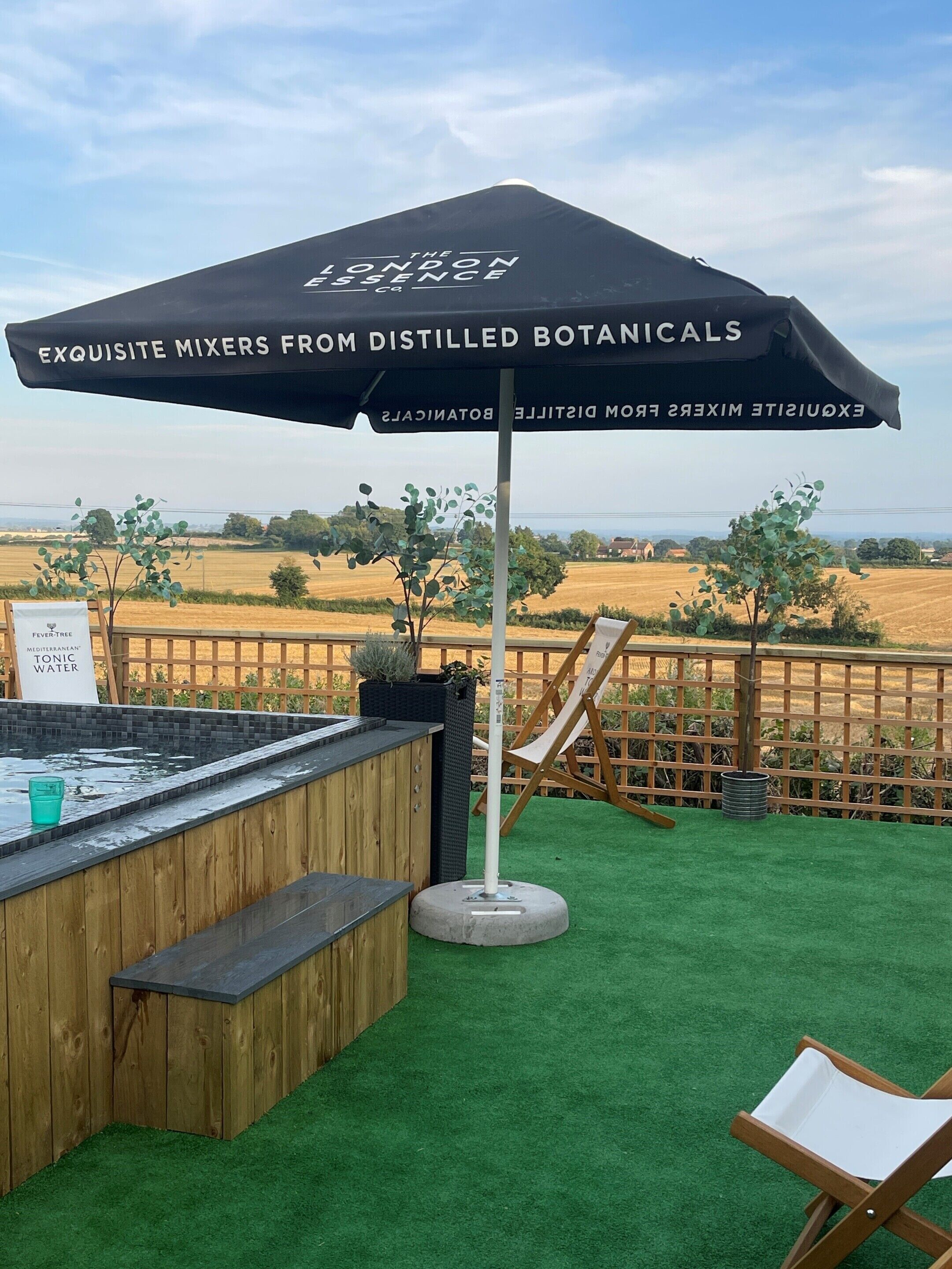 Zona de bar al aire libre con mostrador de madera, césped artificial verde y una sombrilla negra con texto.