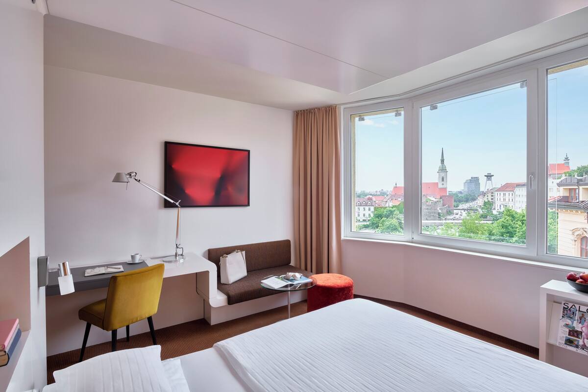 Une chambre d’hôtel comprenant un lit, un bureau avec une chaise, un canapé, une petite table, un téléviseur et une fenêtre donnant sur la ville.