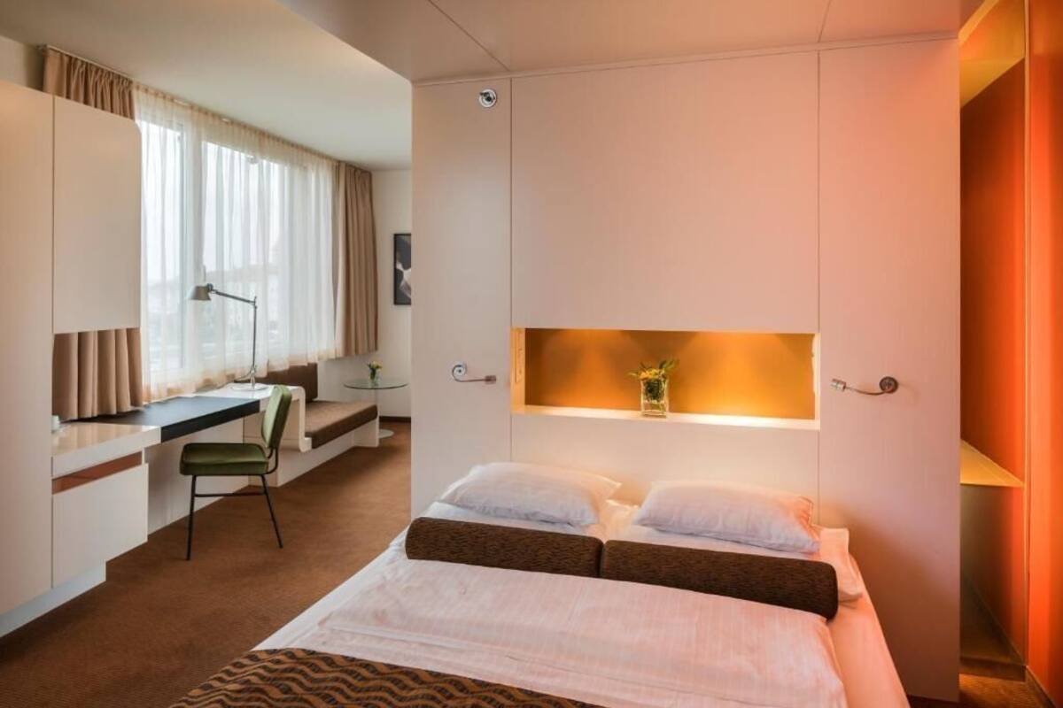 Une chambre d’hôtel avec un lit, un bureau et une chaise. Il y a une fenêtre avec des rideaux, une table de chevet avec une lampe et une petite étagère avec une plante.