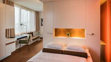 Habitación de hotel con cama, escritorio y silla. Se ve una ventana con cortinas, una mesita de noche con lámpara y un estante pequeño con una planta.