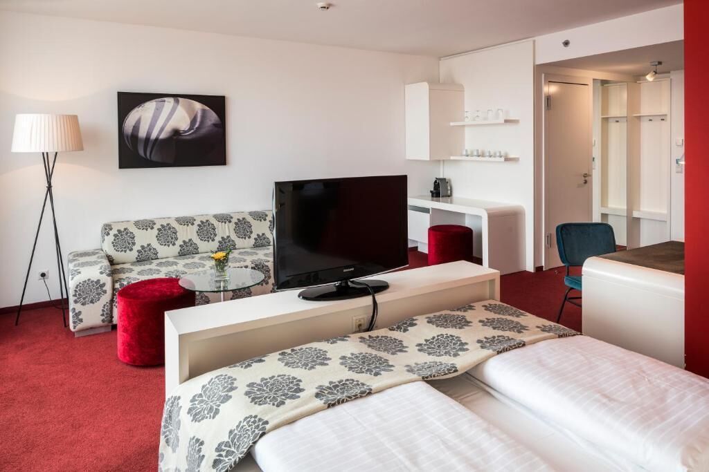 Una habitación de hotel moderna con sofá, televisor, cama, una mesita y un reposapiés rojo.