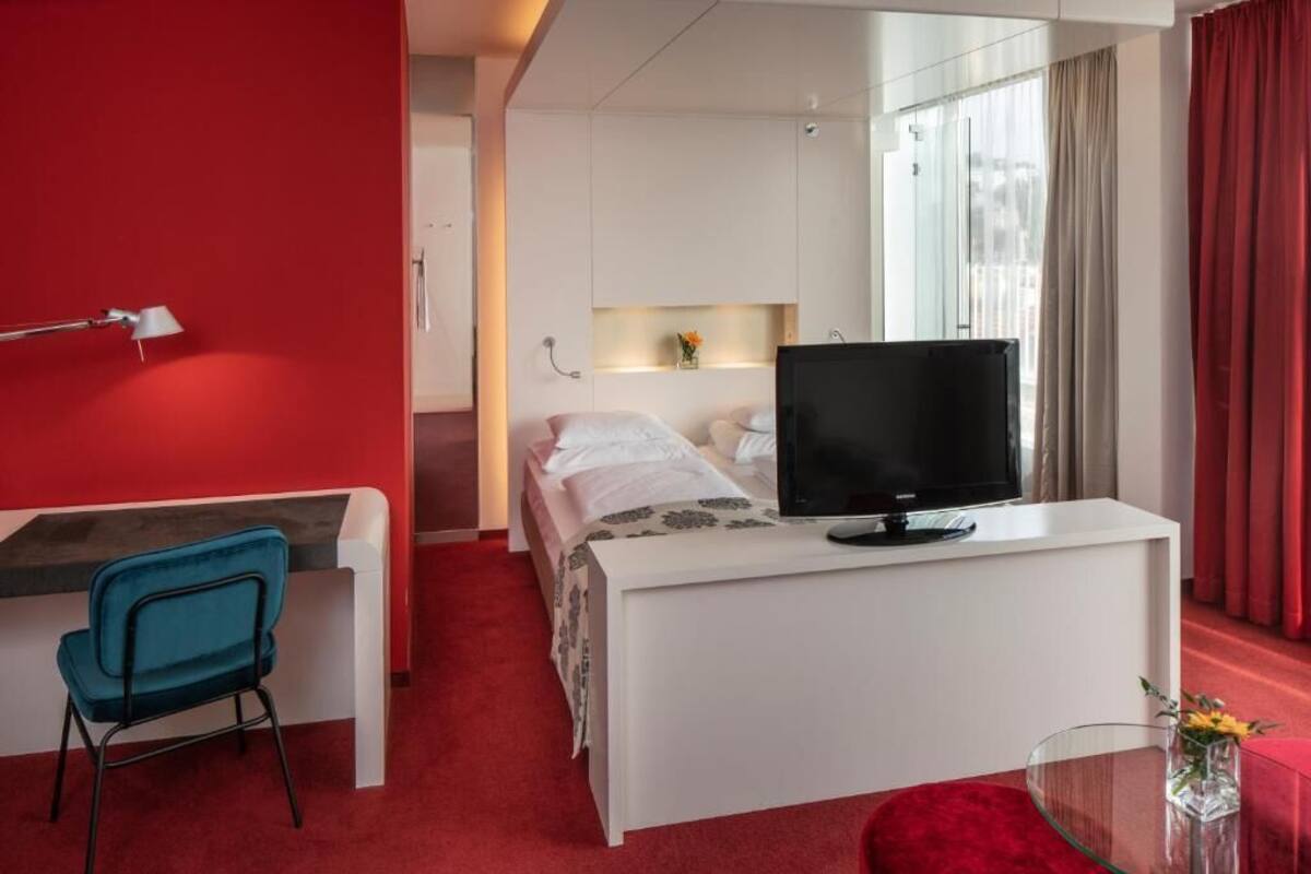 Une chambre d’hôtel moderne avec un lit, un bureau doté d’une télévision, une chaise et une moquette rouge.
