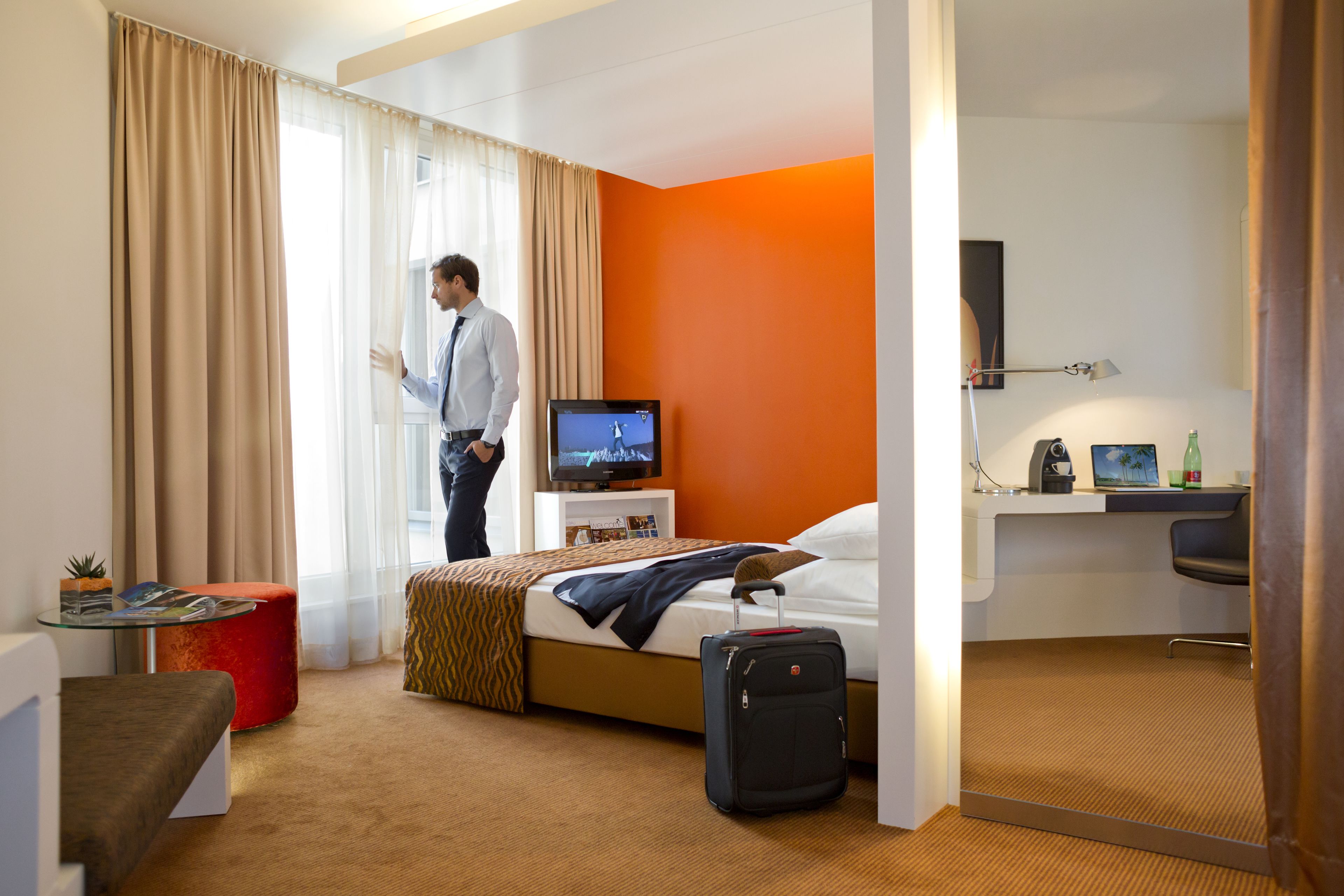 A hotel room with a bed, a desk, a television, a suitcase, and a window with curtains.