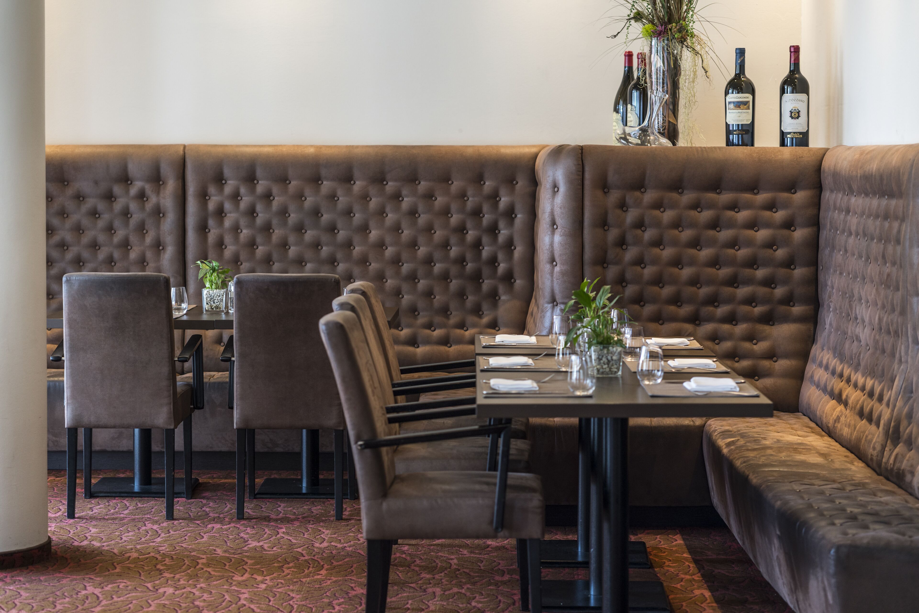 Un restaurante con una banqueta tapizada de color marrón, una mesa dispuesta para dos personas y una zona de bar con botellas.