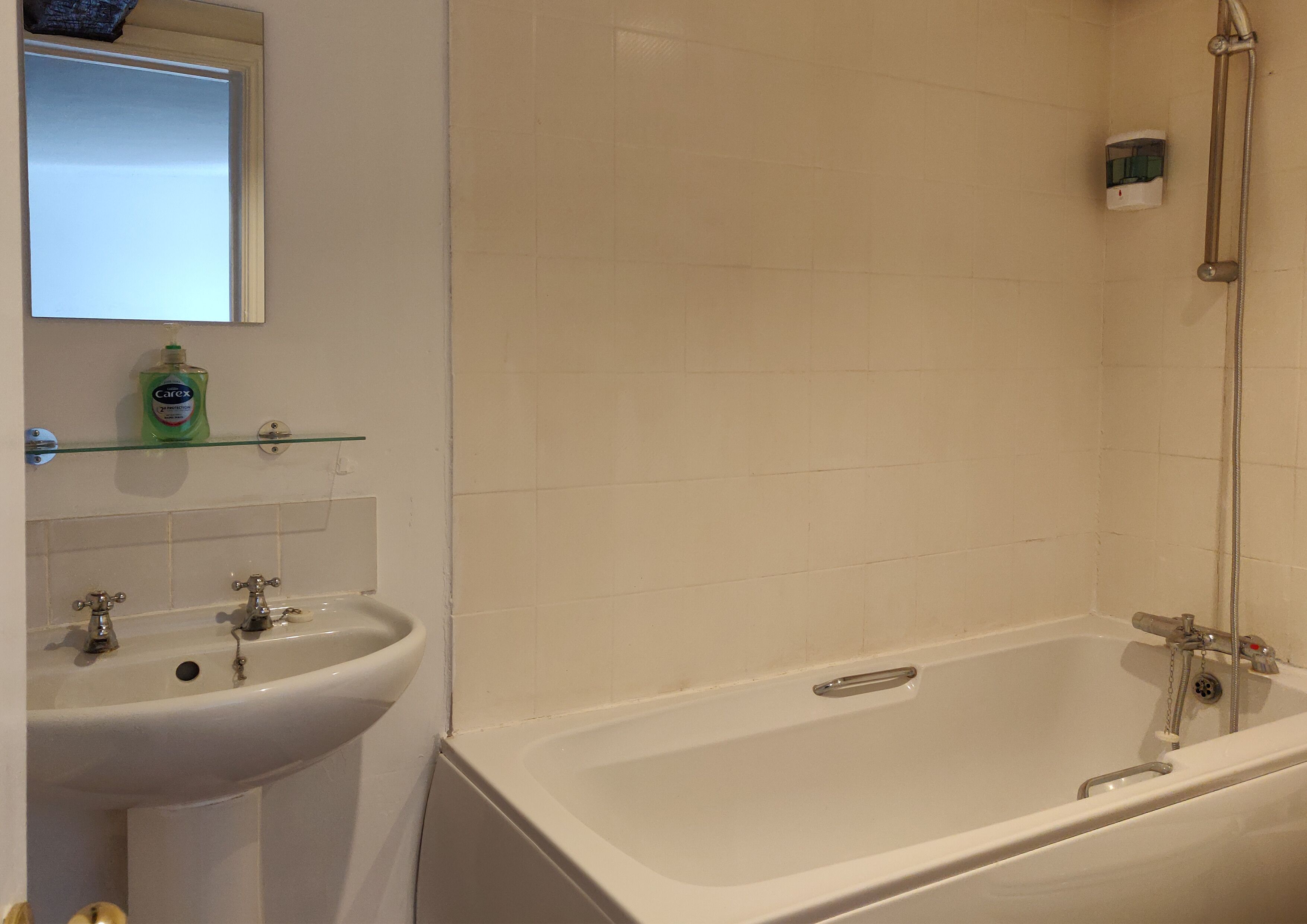 A bathroom with a white sink, two faucets, a glass shelf with a bottle, and a bathtub with a showerhead.