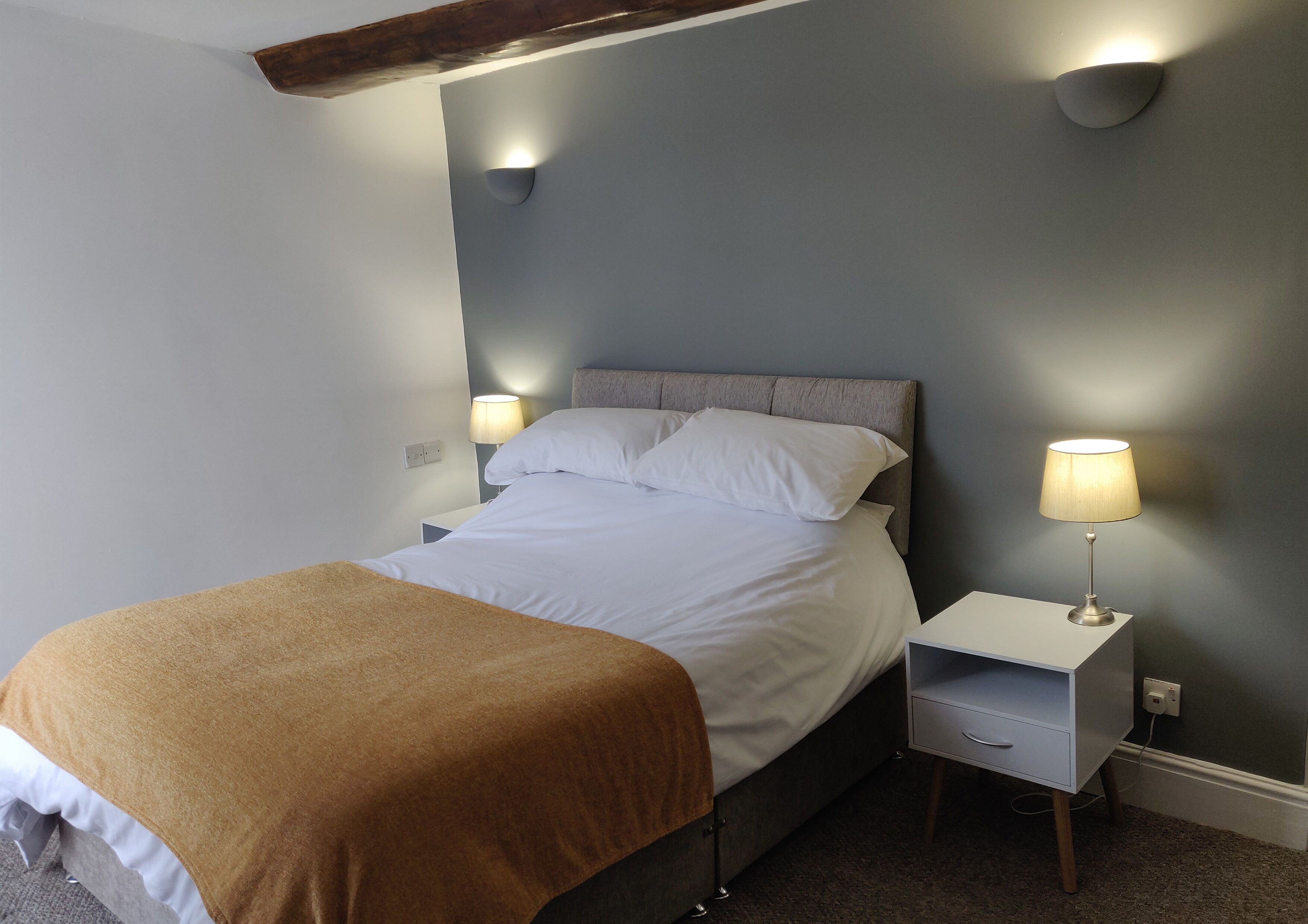 A neatly made bed with a brown blanket, a bedside table with a lamp, and a wall-mounted light fixture.