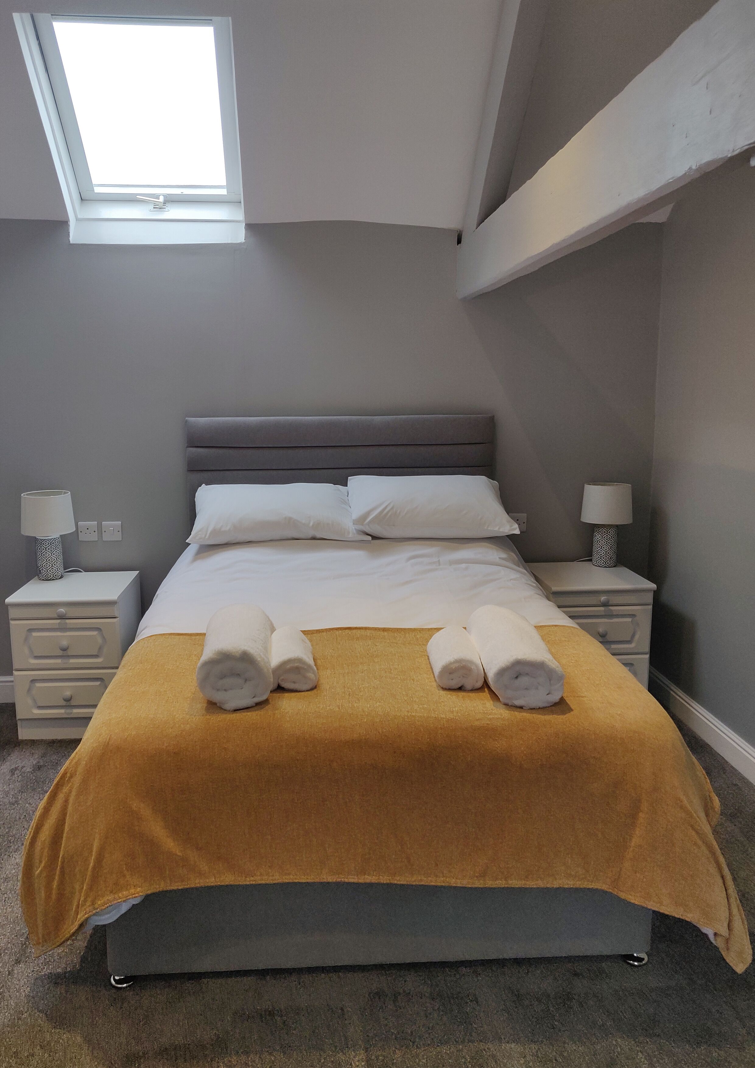 A neatly made bed with a grey headboard, two bedside tables with lamps, a skylight above, and a mustard-colored throw blanket.