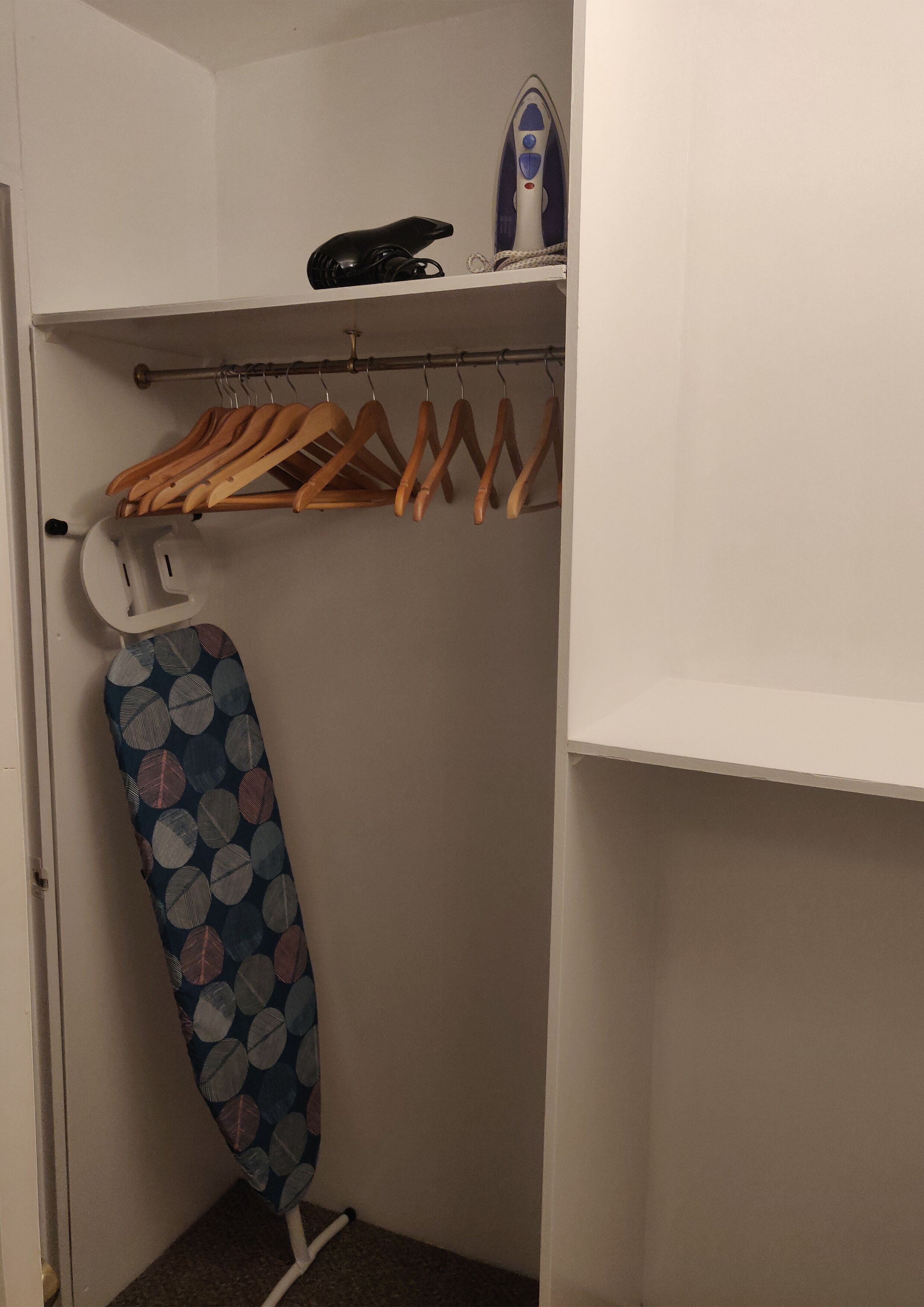 A closet with a hanging rack, an ironing board, and a shelf with a telephone and an iron.