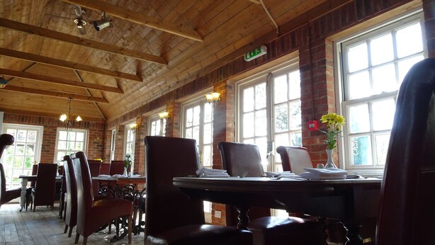 Un restaurante con vigas de madera en el techo, paredes de ladrillo y ventanas grandes. Las mesas están dispuestas con platos y cubiertos.