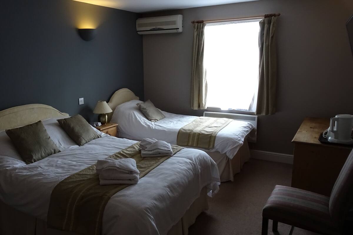 A hotel room with two beds, a nightstand with a lamp, an air conditioning unit, and a window with curtains.