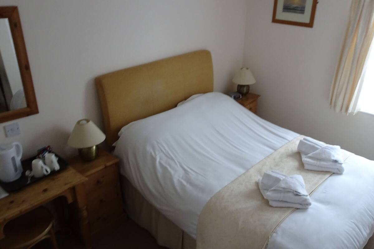 A neatly made bed with white linens, a wooden headboard, and two bedside lamps. There is a small table with a kettle and a teapot on it.
