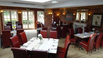 A restaurant with brick walls, wooden tables, and red chairs. There are windows allowing natural light, and a fireplace is visible.