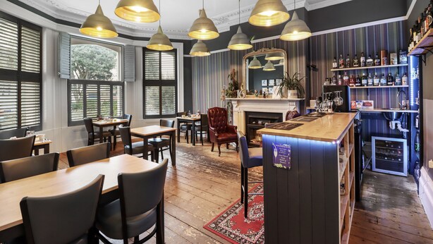 Un restaurante con mesas y sillas de madera, lámparas colgantes, una chimenea y una zona de bar con estantes llenos de botellas.