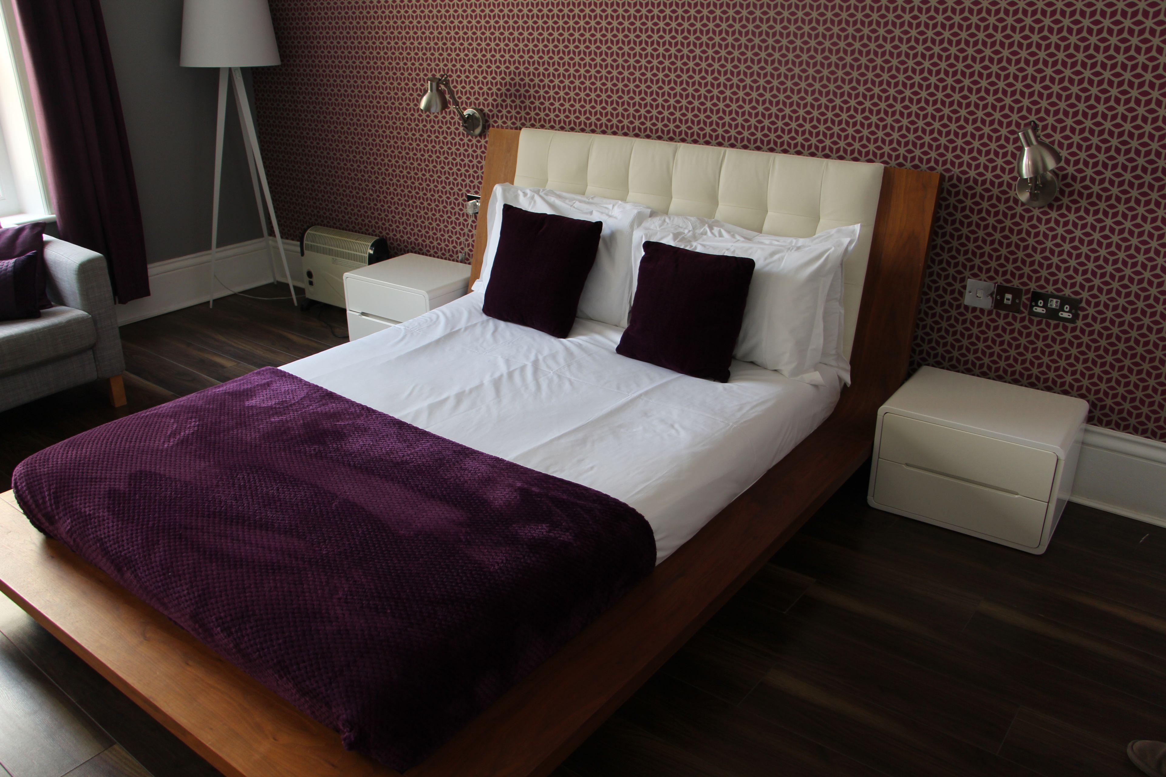 A neatly made bed with a purple throw, a wooden headboard, and white bedding. There are two bedside tables with lamps, a chair, and a patterned wall.
