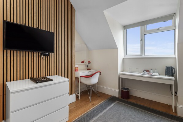 A modern room with a TV mounted on a wooden wall, a white desk with a chair, a small sink, and a window with a view.