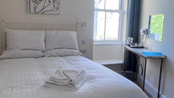 A neatly made bed with white linens and a folded towel, a wall-mounted artwork, a window with curtains, and a small table with a kettle and cups.