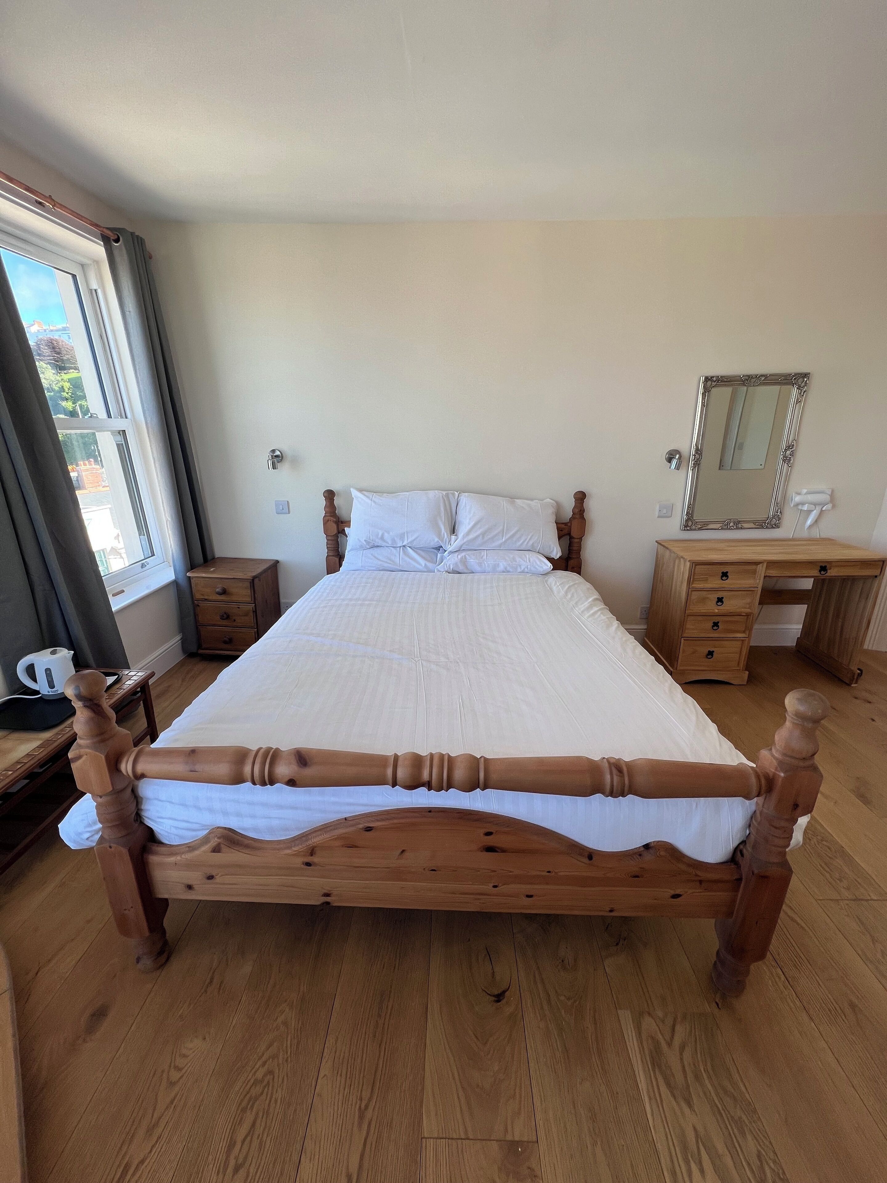 A wooden bed with white bedding, a wooden nightstand, a wooden dresser with drawers, a mirror, and a window with curtains.