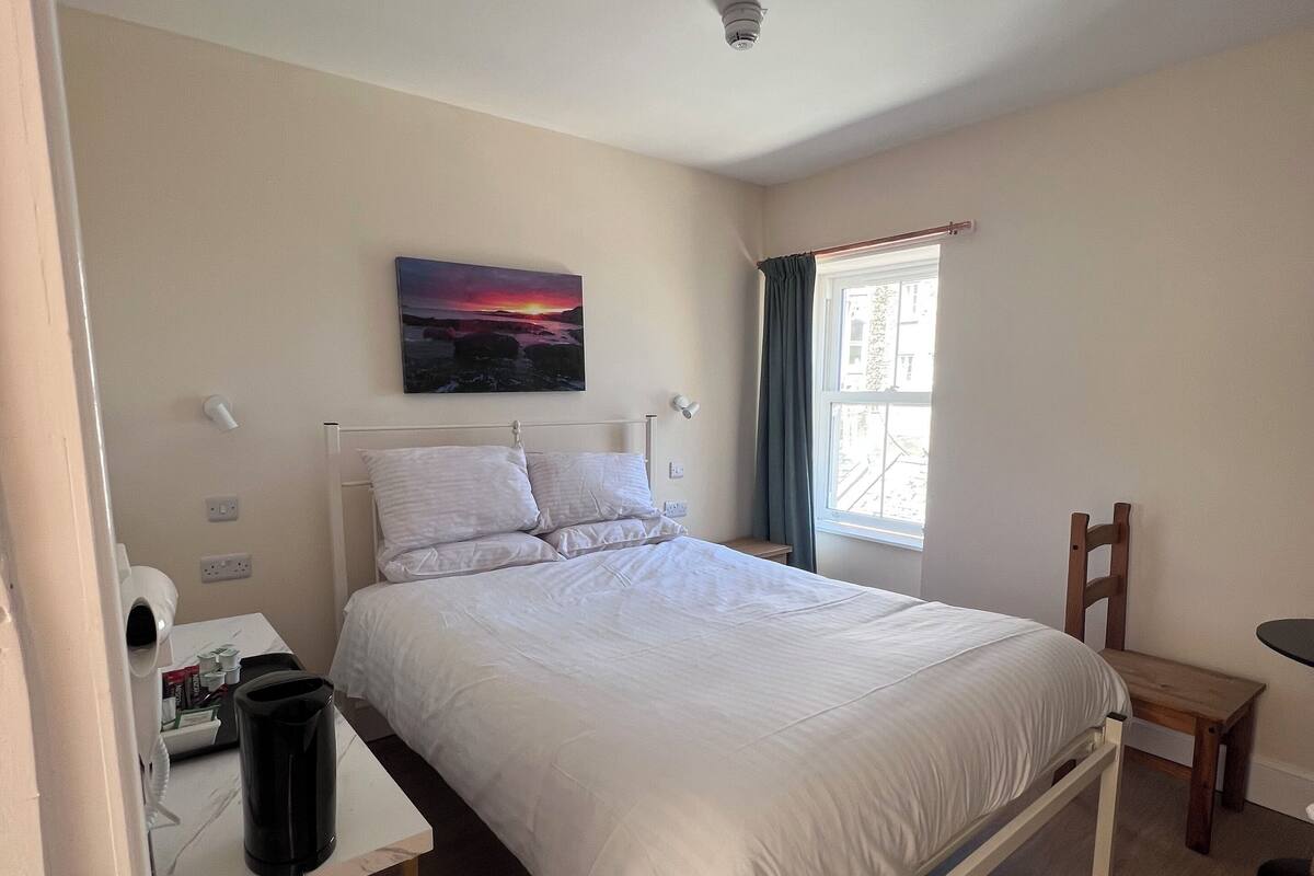 A bedroom with a bed, a desk with a hairdryer, a chair, a window with curtains, and a painting on the wall.