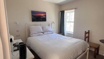 A bedroom with a bed, a desk with a hairdryer, a chair, a window with curtains, and a painting on the wall.