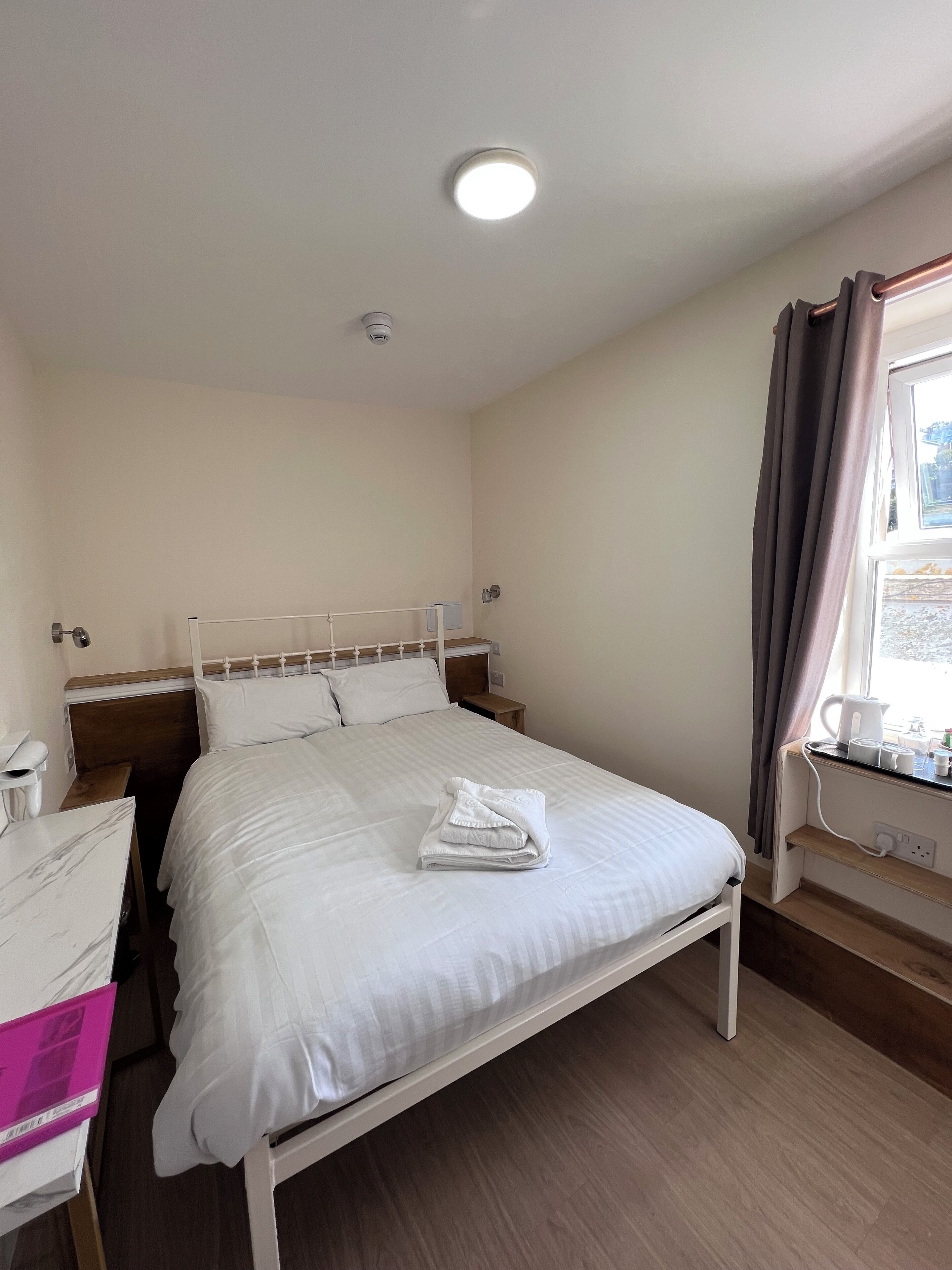A neatly made bed with white linens, a bedside table with a lamp, and a window with curtains.