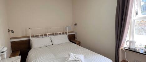A neatly made bed with white linens, a bedside table with a lamp, and a window with curtains.