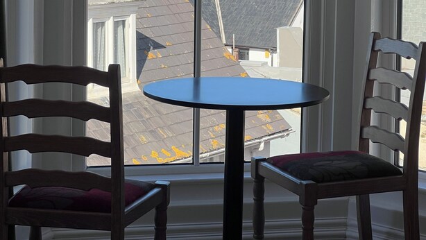 A room with a large window offering a view of the sea and buildings. There is a round table with two chairs. - The Harbour Lights (Ilfracombe)