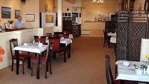 A restaurant with tables and chairs, a counter with a person behind it, and a decorative wall with a clock. - The Harbour Lights (Ilfracombe)