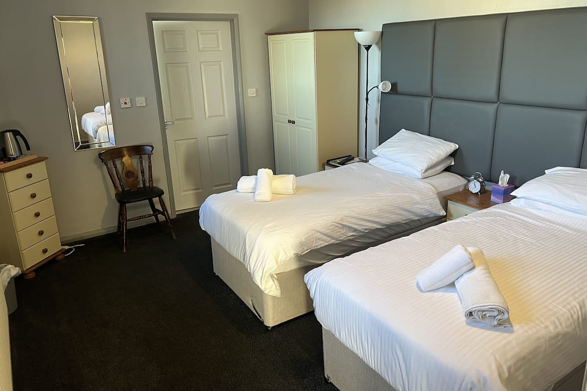 Quarto de hotel com duas camas, uma poltrona, cômoda, espelho e mesa de cabeceira com abajur.