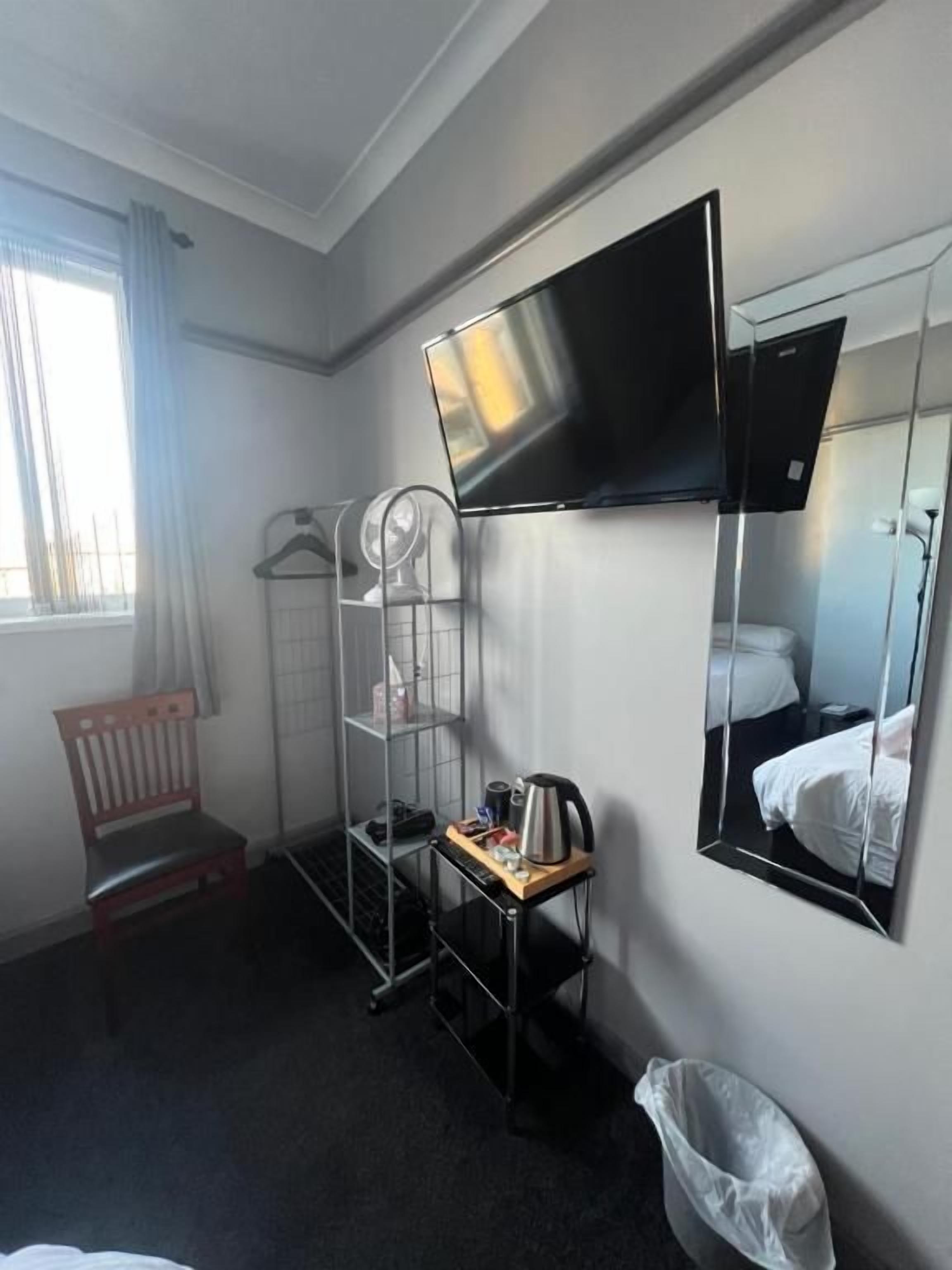 Ein Hotelzimmer mit Bett, Wand-Fernseher, kleinem Tisch mit Wasserkocher, Stuhl, Ablagefach mit Kleiderhaken und Mülleimer.